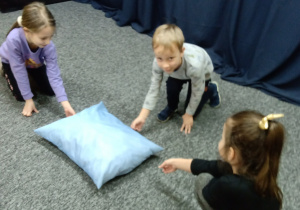 Troje dzieci klęczy na podłodze wokół niebieskiej poduszki.
